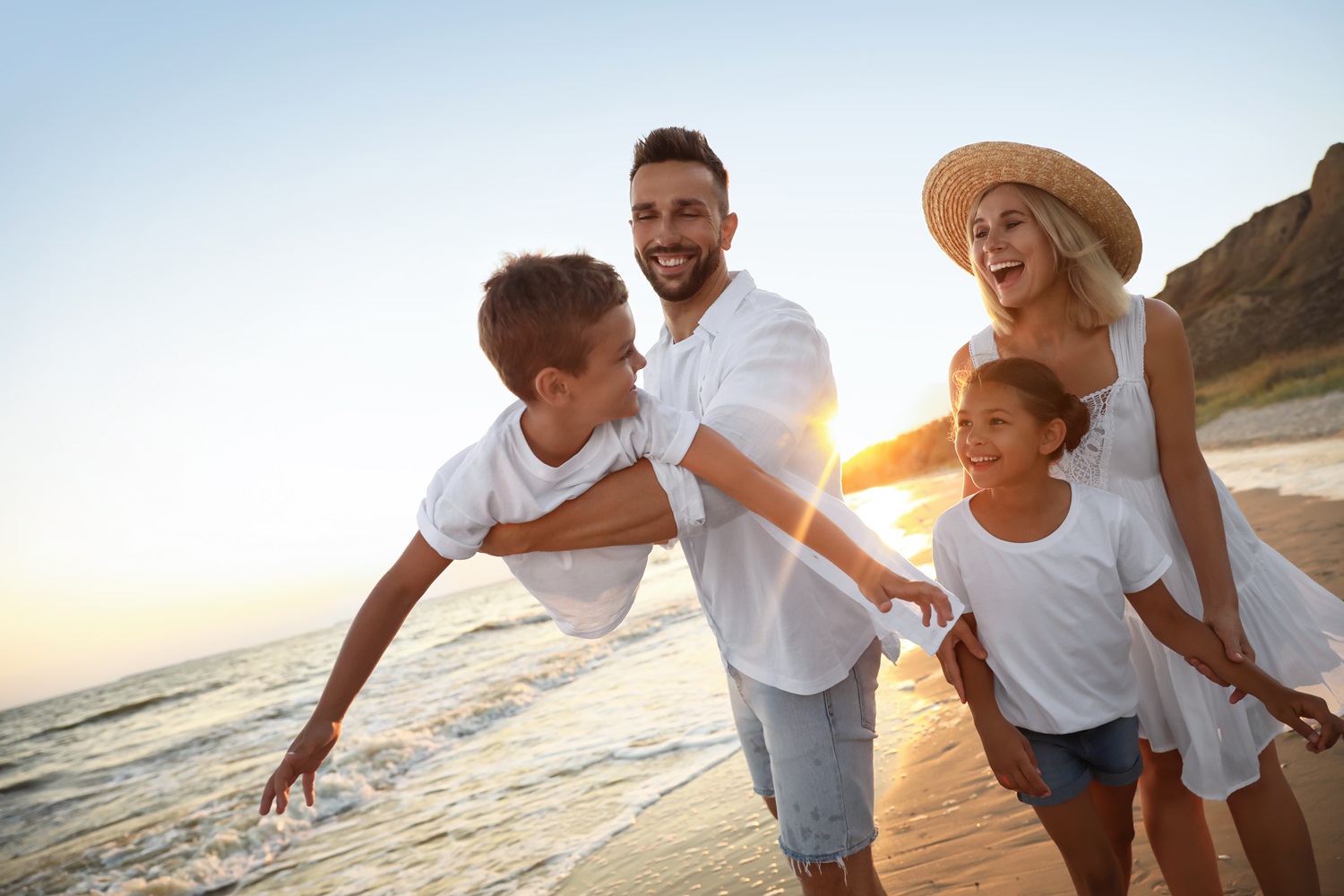 A family of four enjoys walking along the beach at sunset, with the father playfully lifting his son - South Bay Endodontics