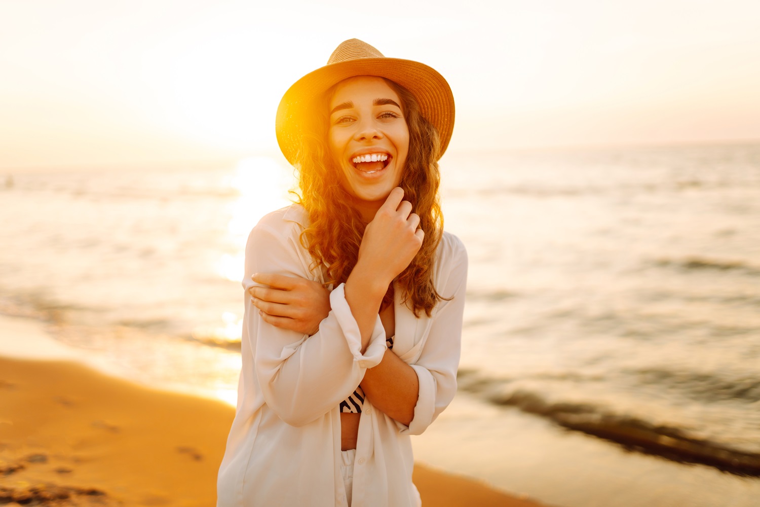 A woman smiling joyfully at the beach during sunset, wearing a straw hat and a light outfit - South Bay Endodontics