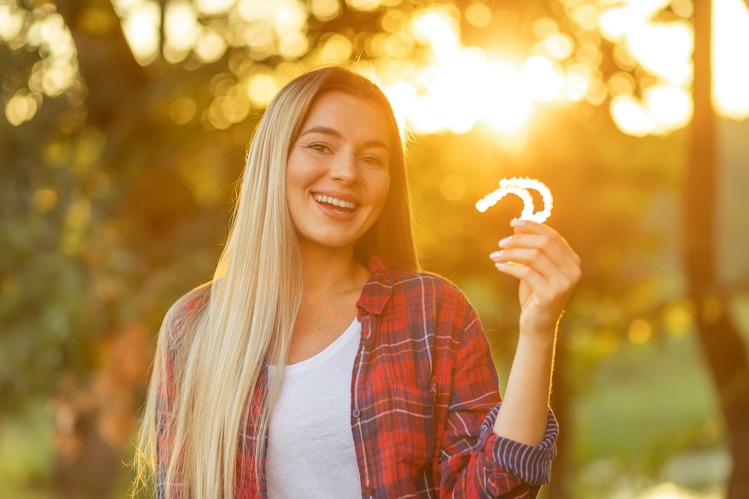 A young woman smiles outdoors while holding a clear dental aligner in her hand - South Bay Endodontics